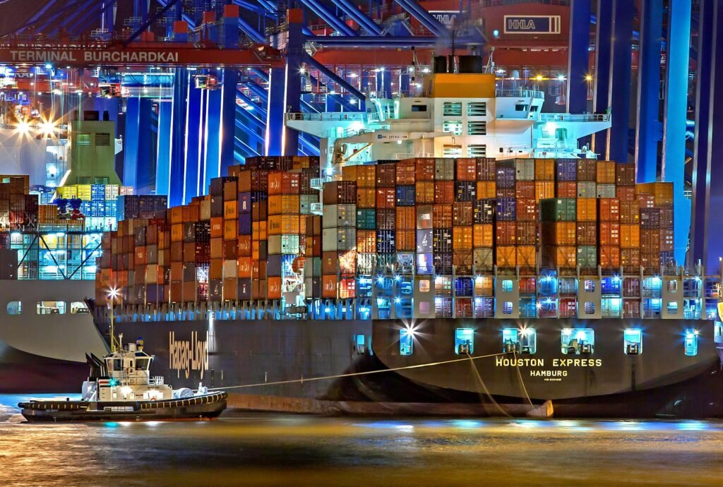 pexels-photo-753331-753331 A brightly lit cargo ship at Hamburg harbor with stacked containers and a tugboat.